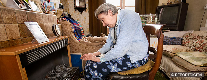 Elderly white woman sitting on chair in front of gasfire to warm hands