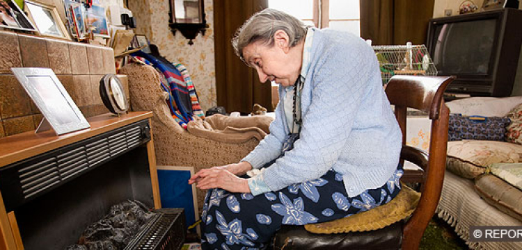 Elderly white woman sitting on chair in front of gasfire to warm hands