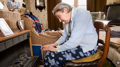 Elderly white woman sitting on chair in front of gasfire to warm hands